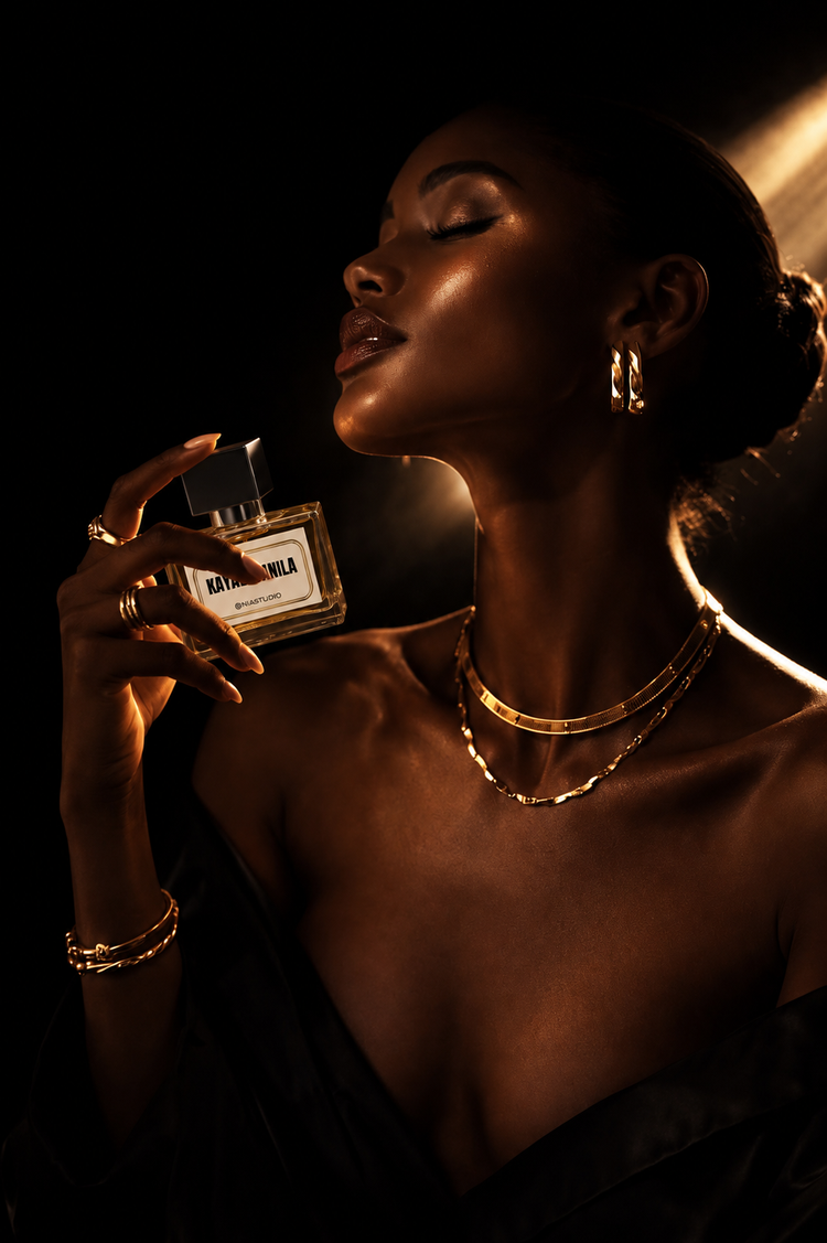 A woman in a black dress holds a rectangular perfume bottle with a clear cap and a white label that reads 'Kayali Vanila'. The scene is lit with dramatic golden lighting against a dark background, showcasing gold jewelry on her.